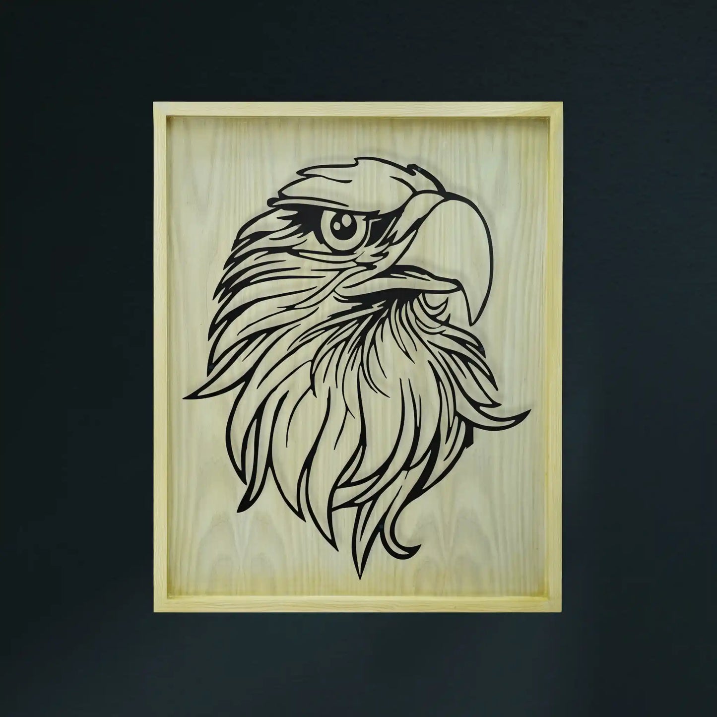A diar wooden framed piece of metal wall art featuring a black line-art illustration of an eagle's head on a light natural wood background, shown against a dark charcoal wall.