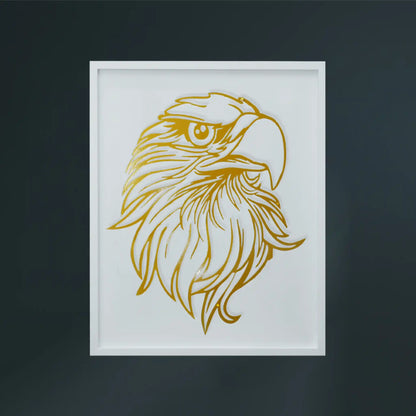 A framed metal wall art featuring a stylized golden eagle head illustration on a white wooden frame, set against a dark gray wall.