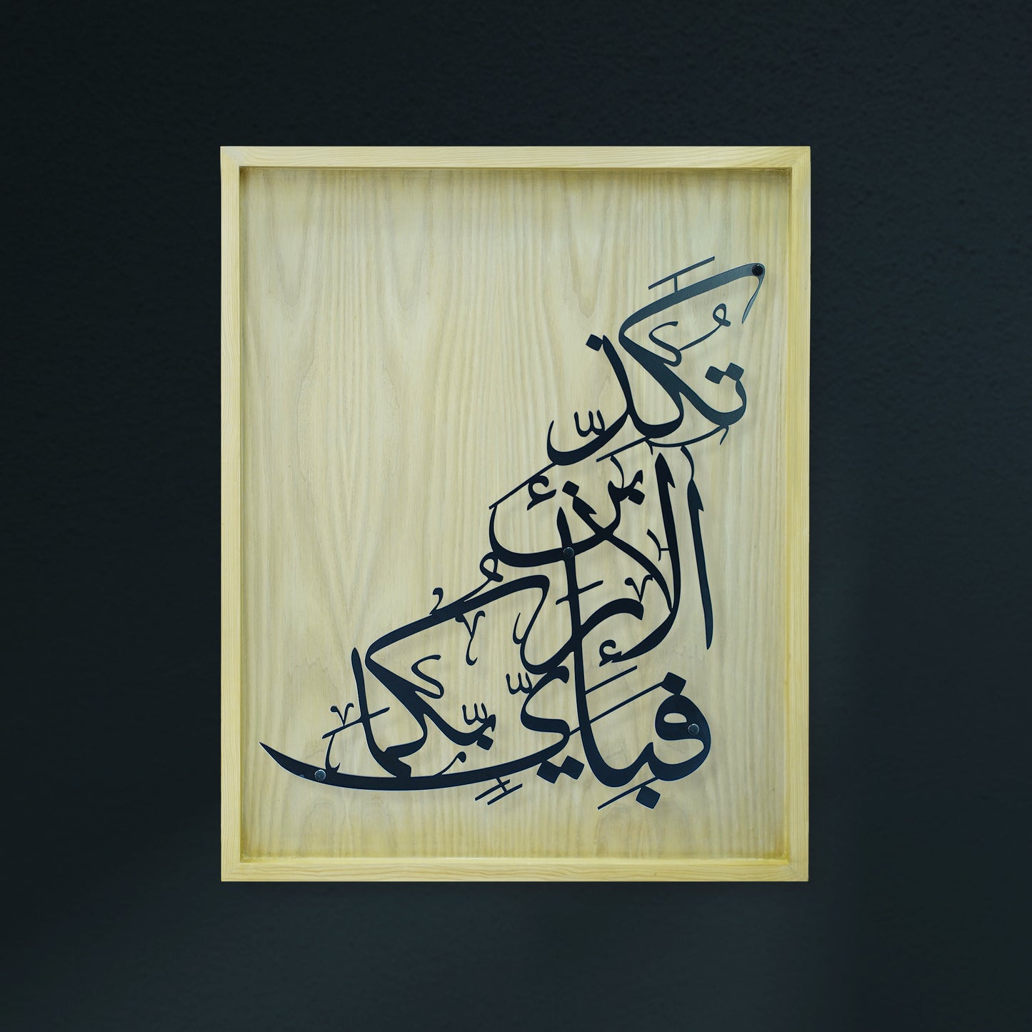 Arabic calligraphy metal wall art in black on a light natural wood panel, mounted in a light wood frame against a dark charcoal background.