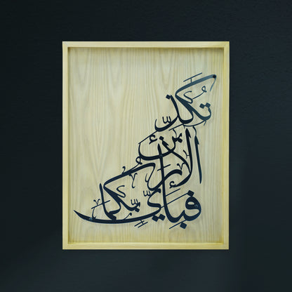 Arabic calligraphy metal wall art in black on a light natural wood panel, mounted in a light wood frame against a dark charcoal background.