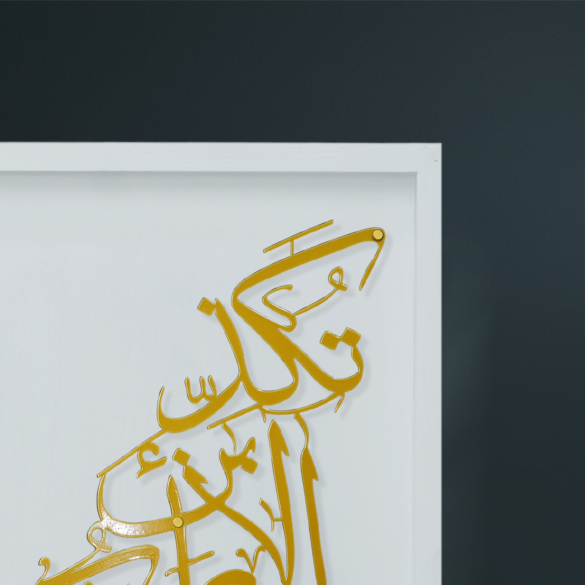 Gold Arabic calligraphy metal wall art mounted on a white wooden frame, shown against a dark charcoal background.