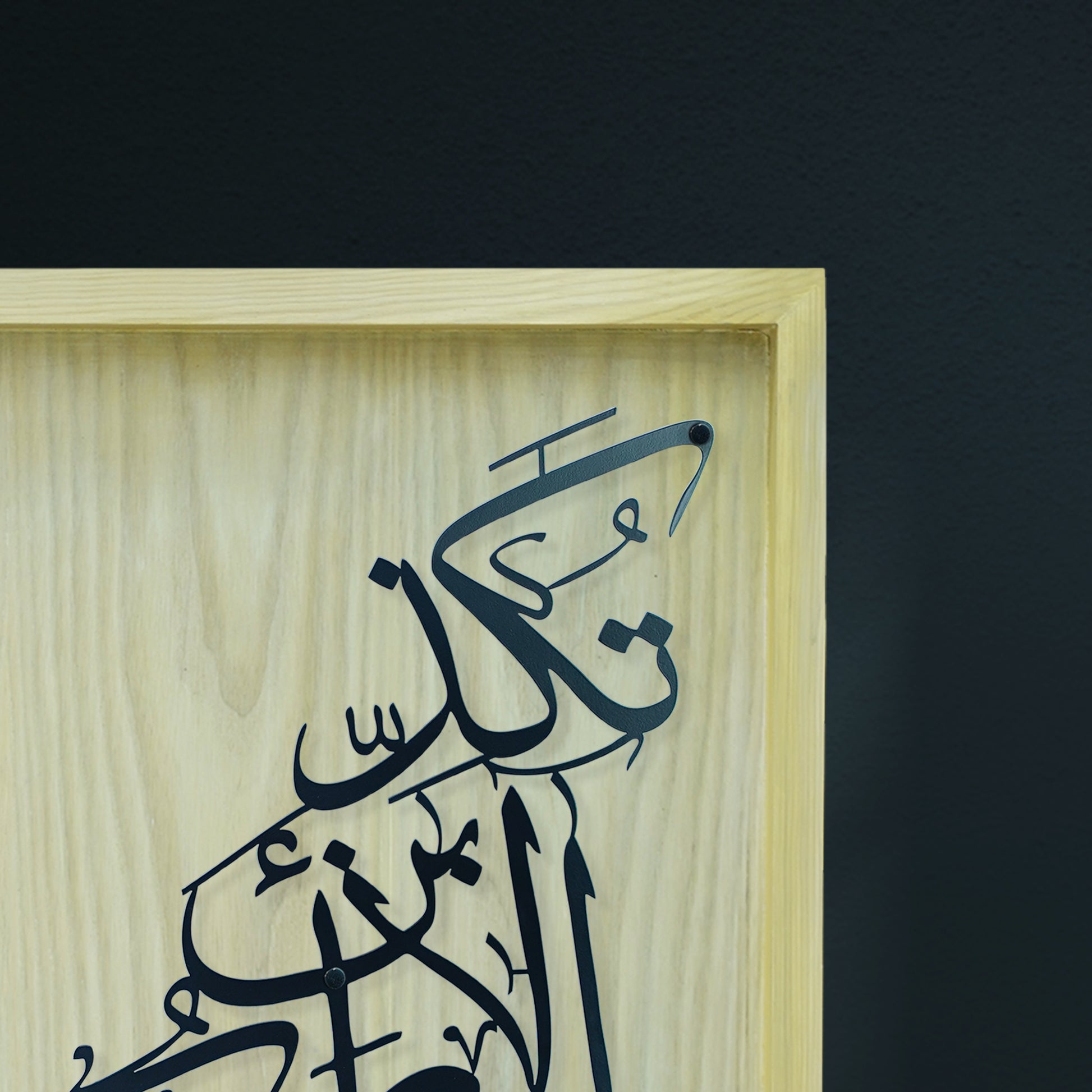 A close-up of a framed piece of Arabic calligraphy on a light natural wood panel, set against a dark charcoal wall.