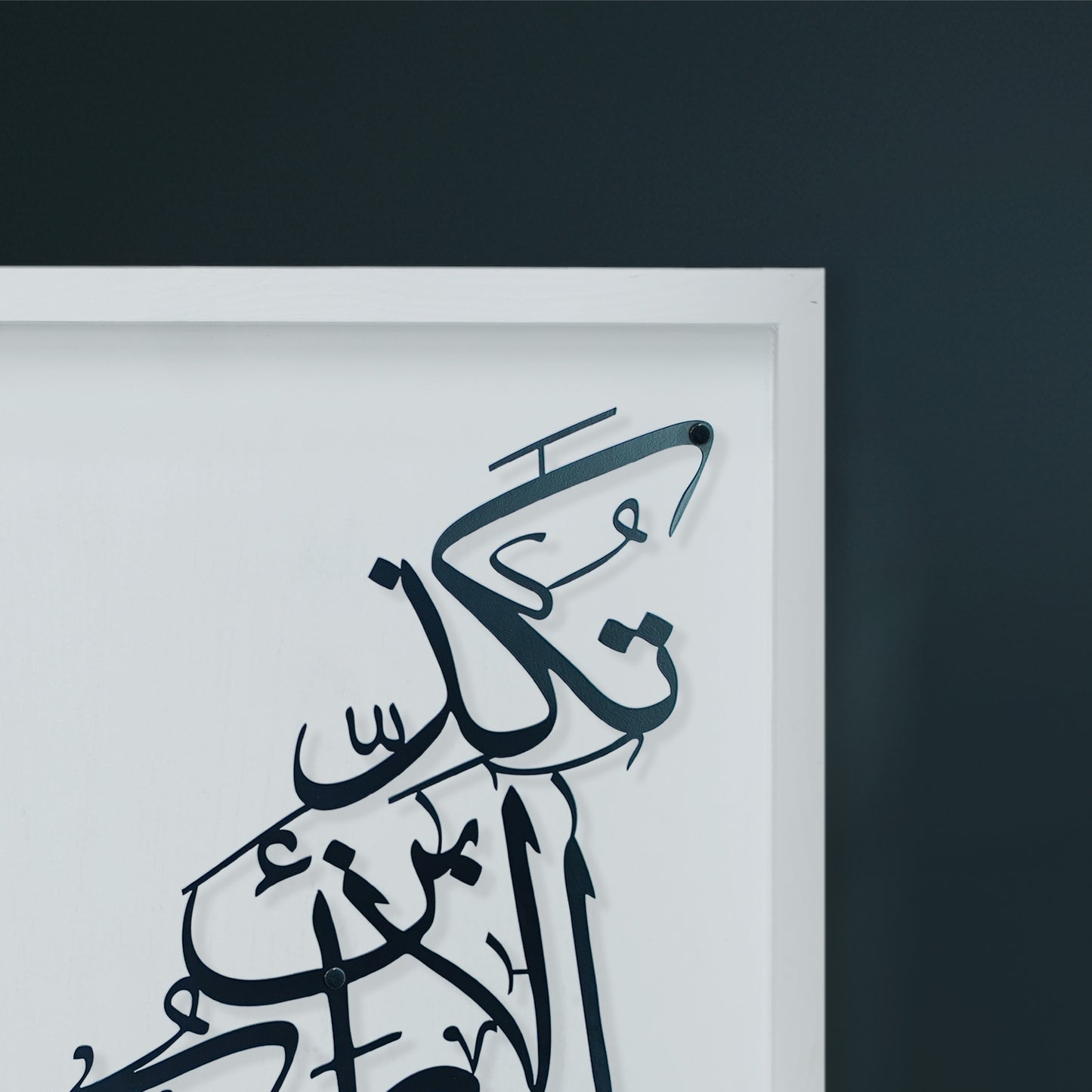 Close-up of a framed Arabic calligraphy metal wall art piece in black on a white wooden, mounted against a dark charcoal wall.