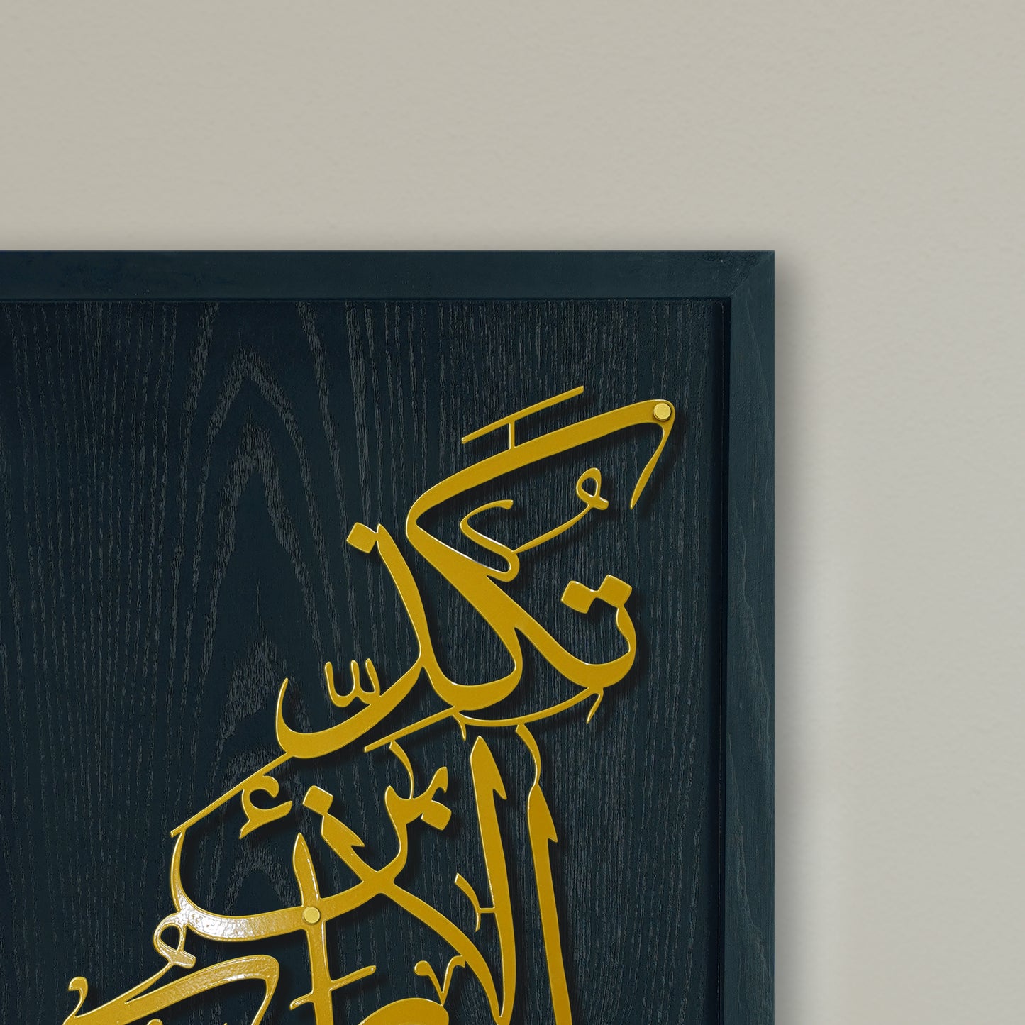 Gold Arabic calligraphy metal wall art mounted on a black wood panel, shown in a close-up against a light beige wall.
