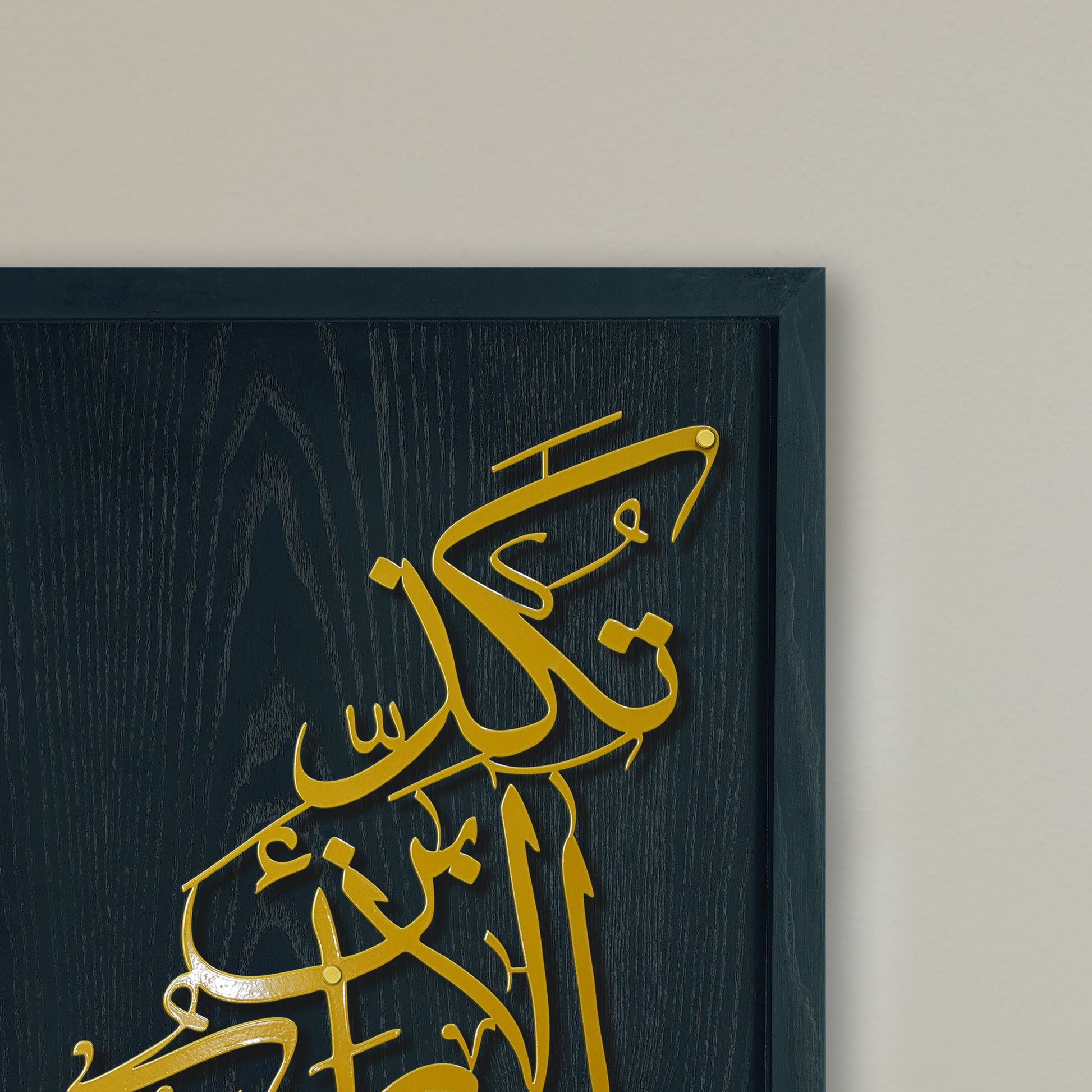 Gold Arabic calligraphy metal wall art mounted on a black wood panel, shown in a close-up against a light beige wall.