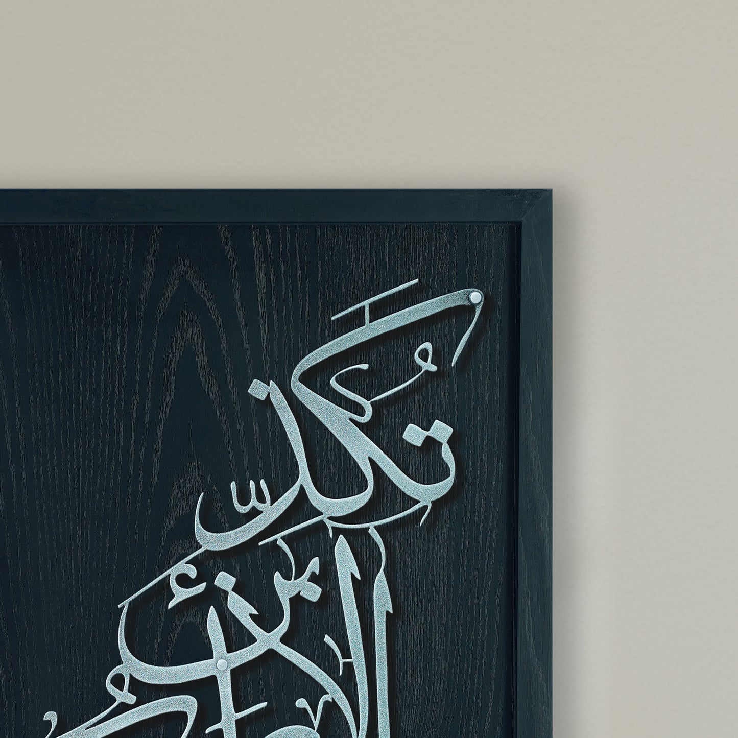 A close-up of framed Arabic calligraphy metal wall art mounted on a light beige wall. The artwork features light silver calligraphy layered over a dark charcoal wood-textured background, enclosed in a dark navy frame.