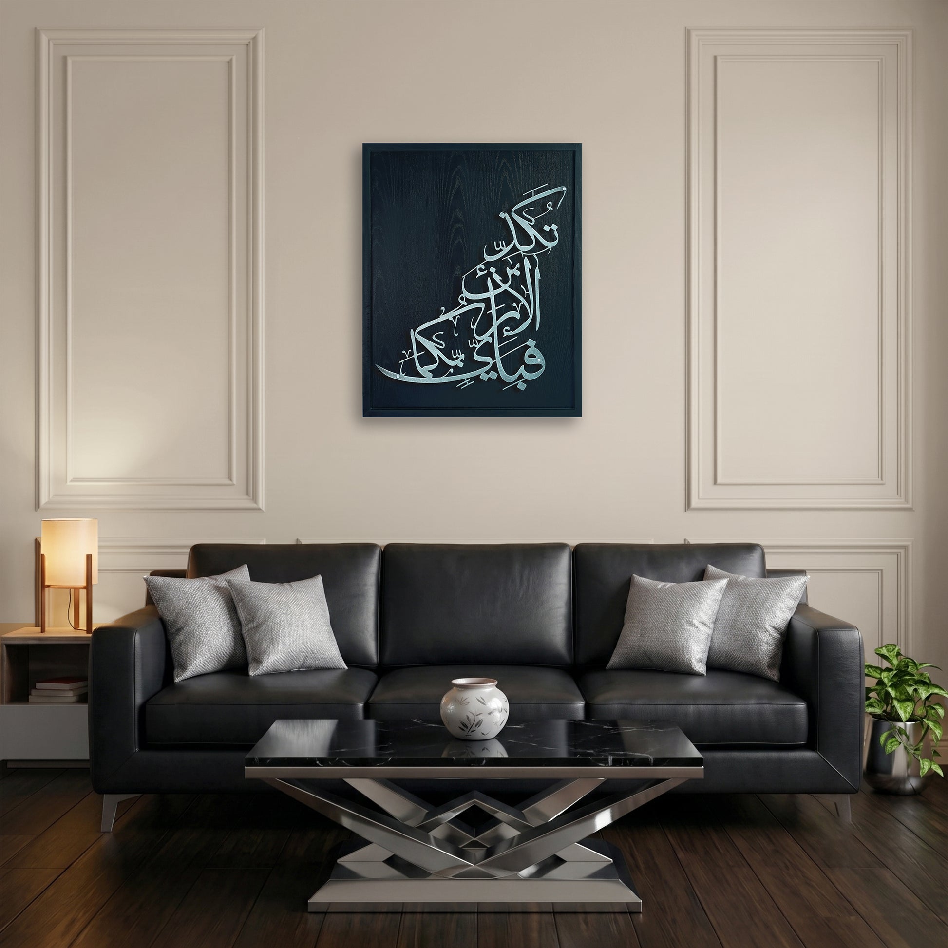 A framed vertical canvas print featuring silver Arabic calligraphy on a black wooden frame, displayed on a beige wall above a black leather sofa in a modern living room setting.