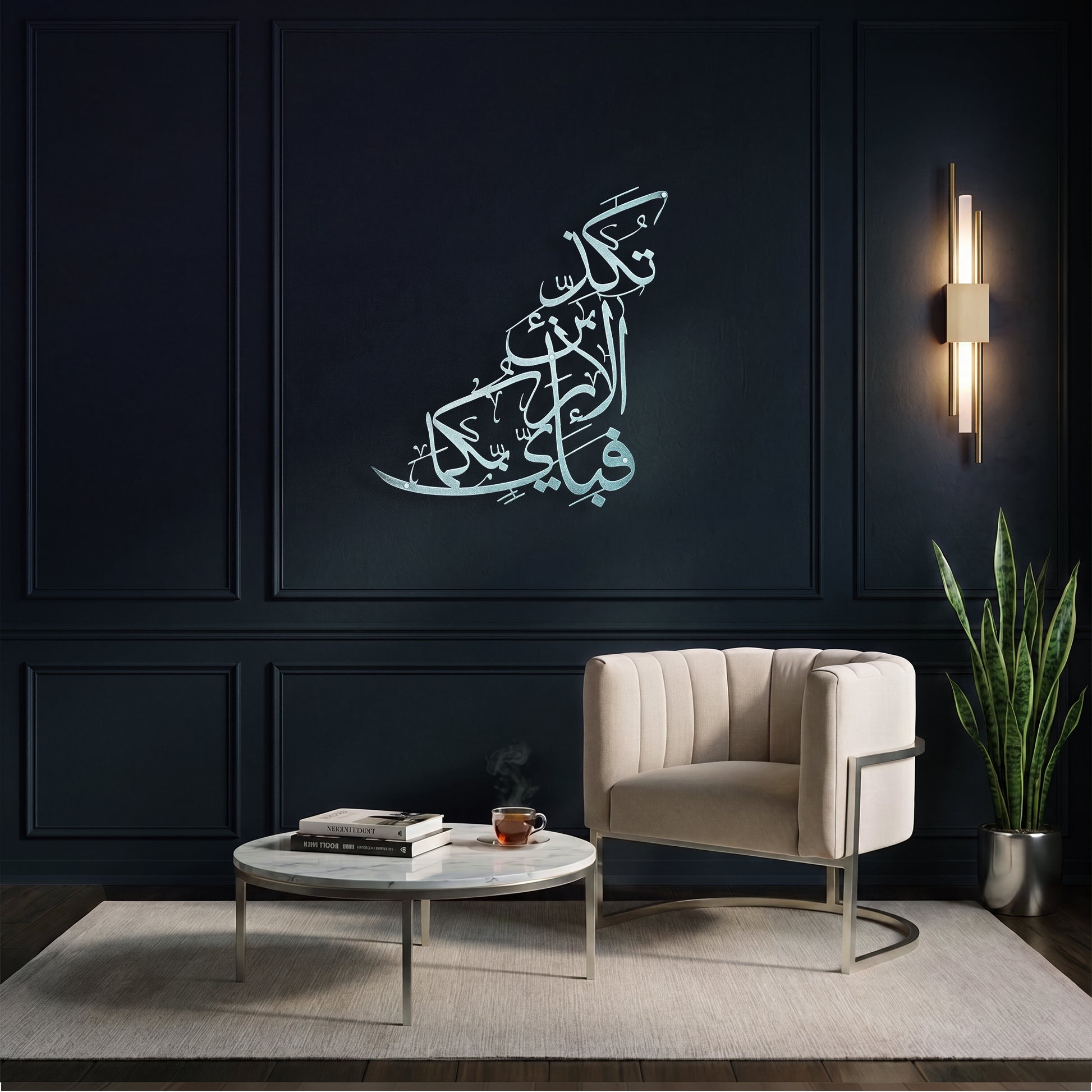 A styled interior scene with a dark navy paneled wall featuring a large silver Arabic calligraphy metal wall art piece above a cream upholstered armchair, a round white coffee table, and a potted snake plant.