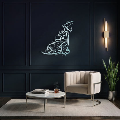 A styled interior scene with a dark navy paneled wall featuring a large silver Arabic calligraphy metal wall art piece above a cream upholstered armchair, a round white coffee table, and a potted snake plant.
