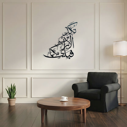 Black Arabic calligraphy metal wall art displayed on a cream paneled wall in a modern living room setting with a dark gray armchair, wooden coffee table, and floor lamp.