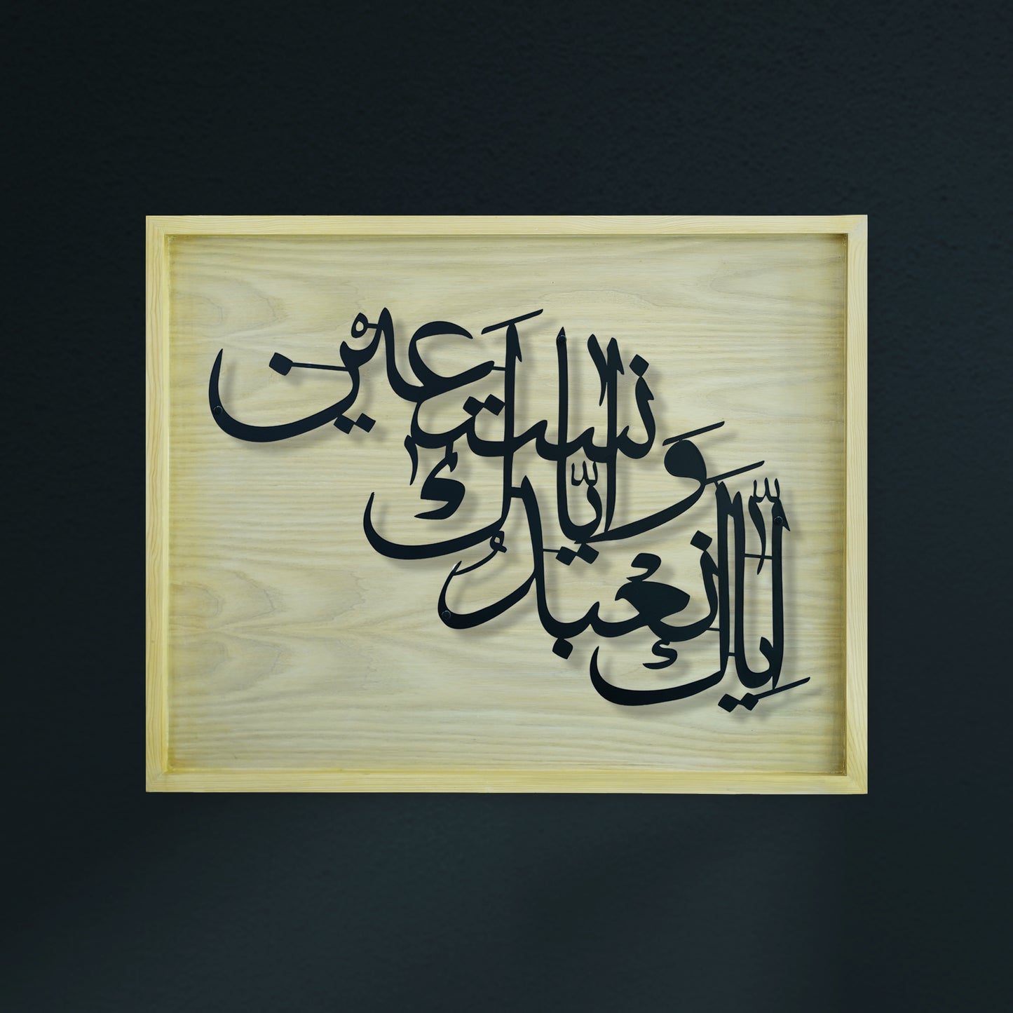 Arabic calligraphy metal wall art in black layered metal over a light natural wood diar frame, shown against a dark charcoal background.