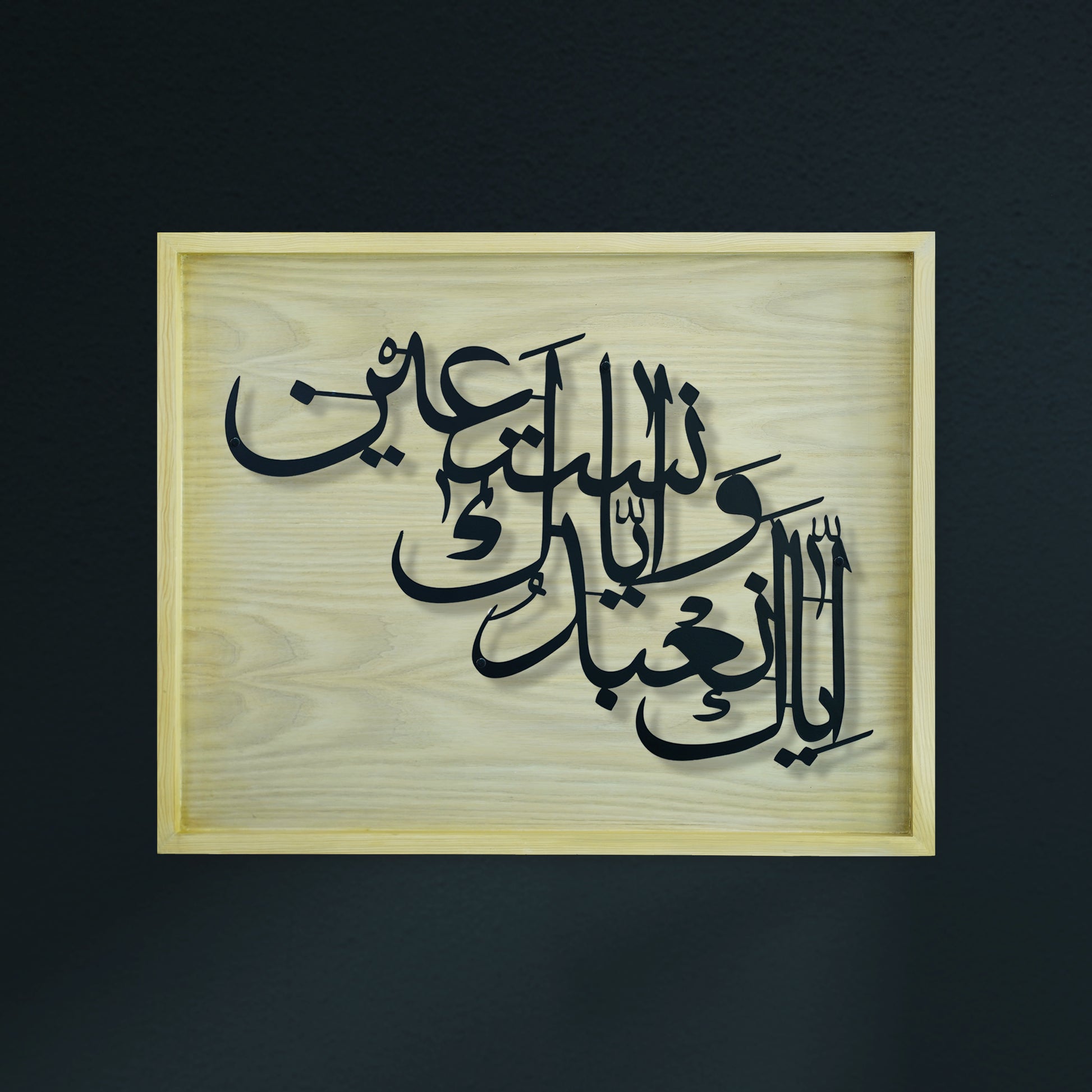 Arabic calligraphy metal wall art in black layered metal over a light natural wood diar frame, shown against a dark charcoal background.