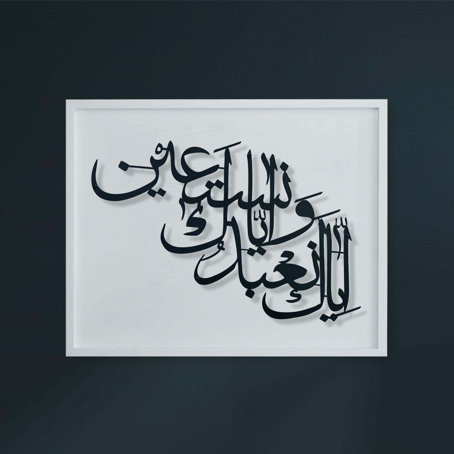 Arabic calligraphy metal wall art in black on a white wooden, shown in a white frame against a dark charcoal wall.
