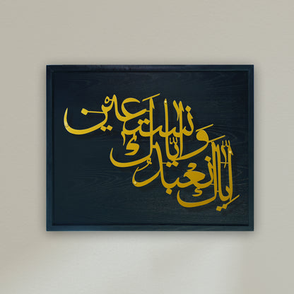 A wooden framed with metal wall art piece featuring elegant gold Arabic calligraphy on a dark black frame, mounted on a light beige wall.