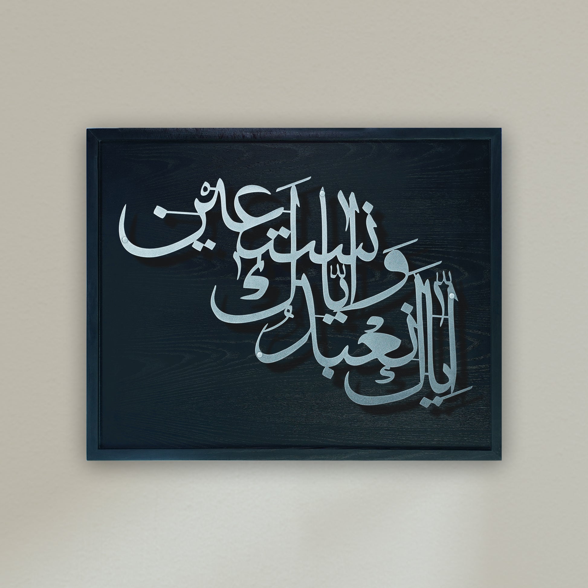 Arabic calligraphy metal wall art in silver on a dark black wooden frame, mounted against a plain beige wall.