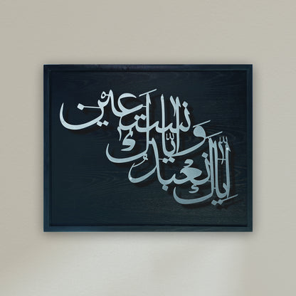 Arabic calligraphy metal wall art in silver on a dark black wooden frame, mounted against a plain beige wall.