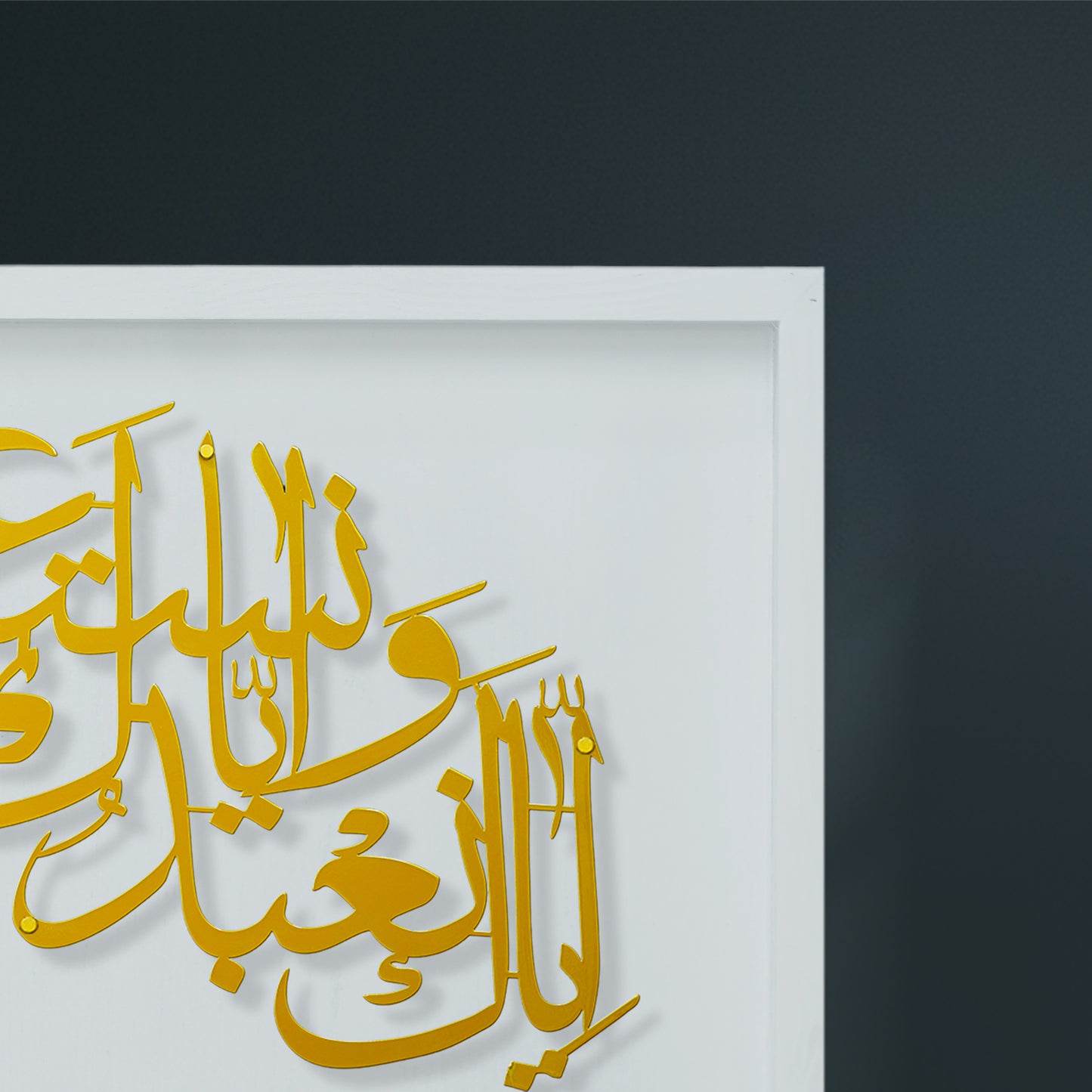Gold laser-cut Arabic calligraphy metal wall art mounted on a white wooden frame, shown against a dark charcoal gray wall.
