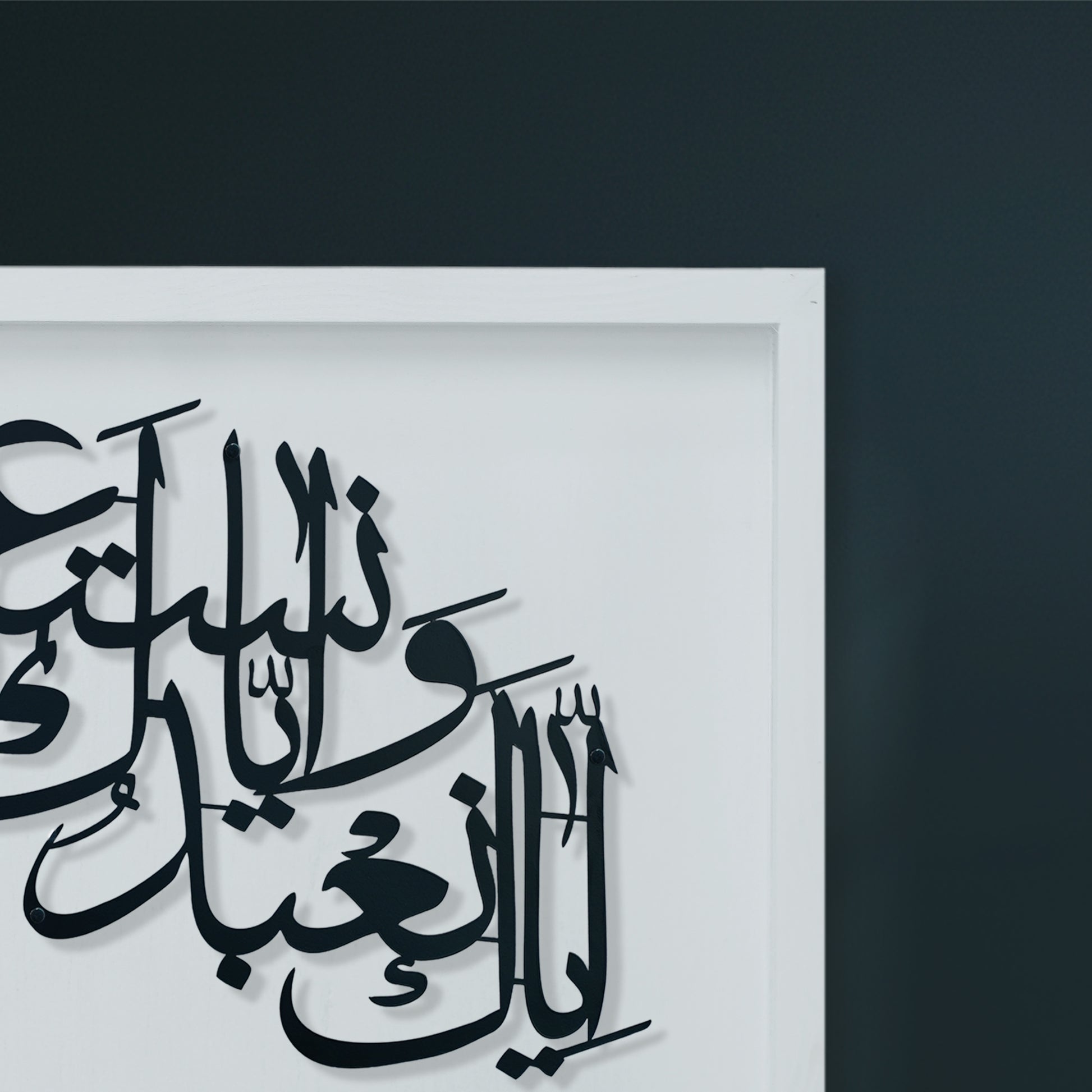 Black Arabic calligraphy metal wall art mounted on a white wooden frame, shown against a dark charcoal wall.