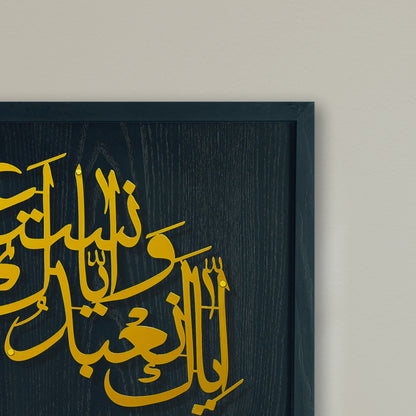 Close-up of a framed metal wall art piece with gold Arabic calligraphy on a dark black wood frame, mounted against a light beige wall.