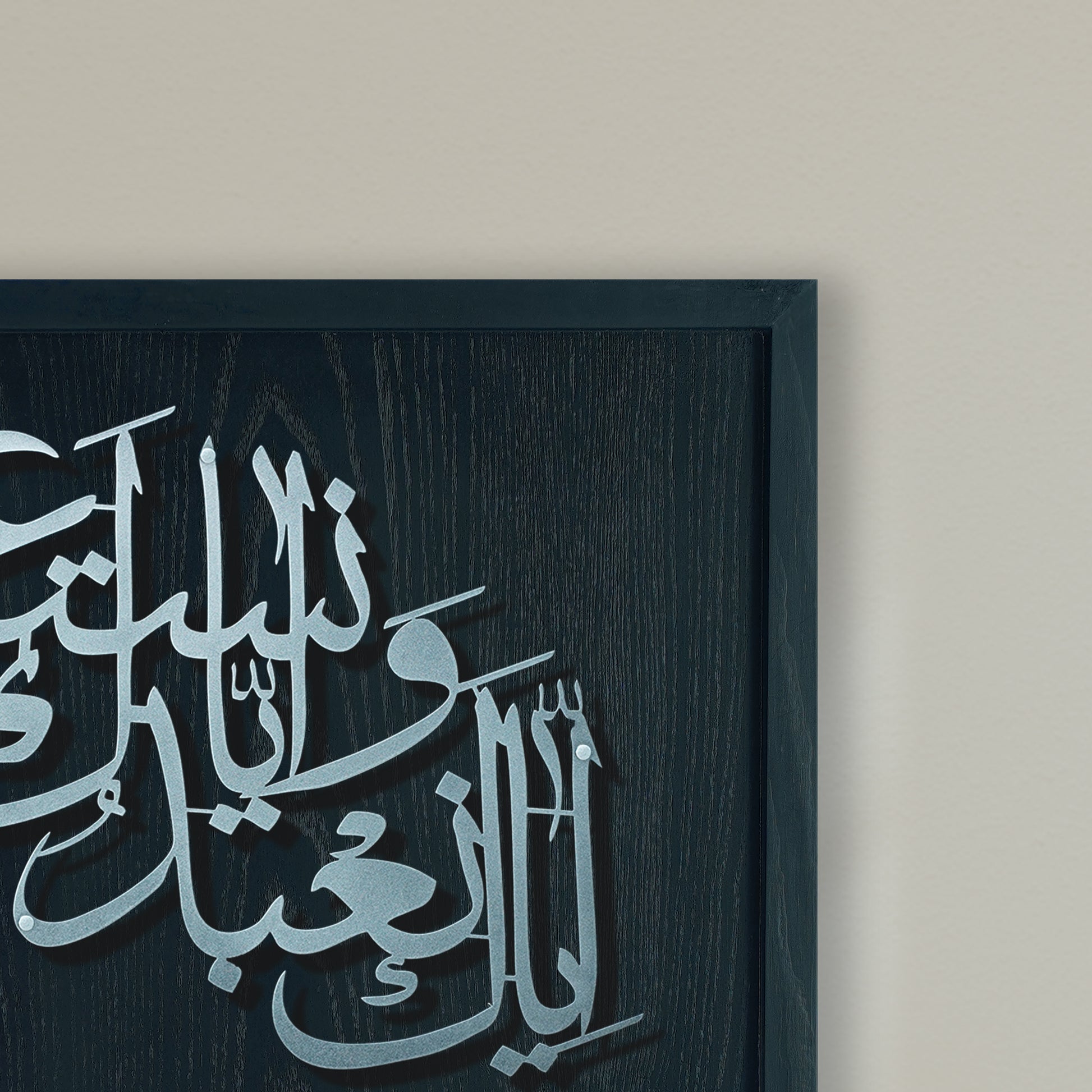 A close-up of framed metal Arabic calligraphy wall art on a dark wood frame, mounted against a light beige wall.