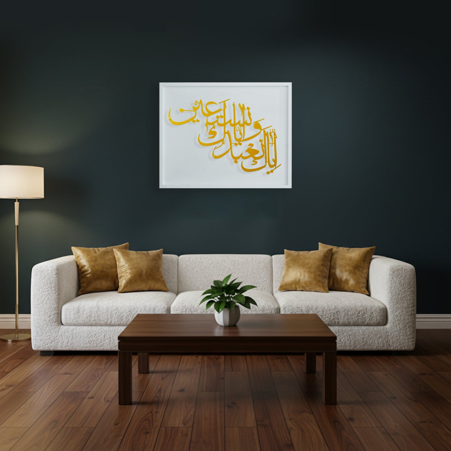 A framed metal wall art with gold Arabic calligraphy on a white wooden frame, displayed above a modern white boucle sofa in a dark living room with a wooden coffee table and wood floor.