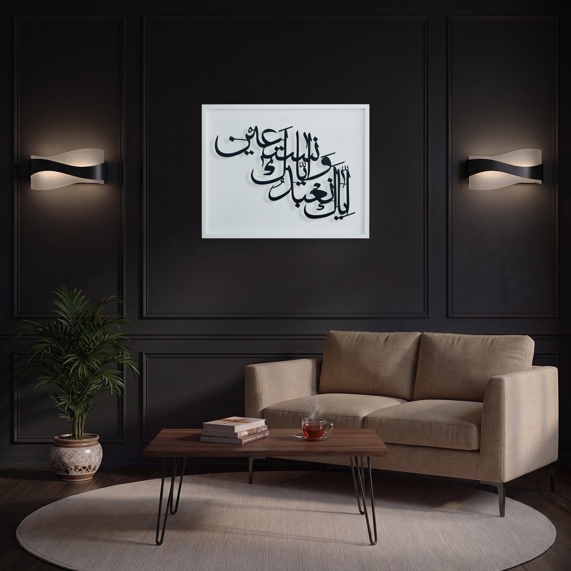A framed black Arabic calligraphy metal wall art on a white wooden frame is displayed in a modern living room setting above a beige sofa, with black paneled walls and wall sconces in the background.