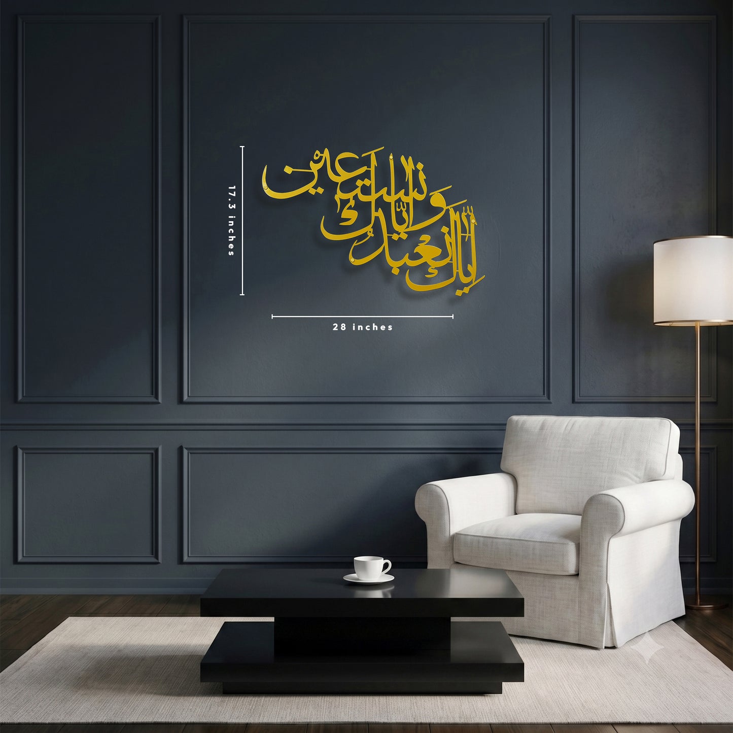 Gold Arabic calligraphy metal wall art with dimension markings 17.3 tall and 28 inch width, displayed on a dark paneled wall above a white armchair and black coffee table.