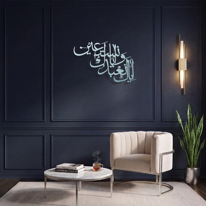 A modern living room setting featuring a dark navy blue paneled wall with elegant Arabic calligraphy metal wall art. In front of the wall is a cream-colored tufted armchair, a round white coffee table with books and a cup, and a tall green potted snake plant next to a vertical wall sconce.