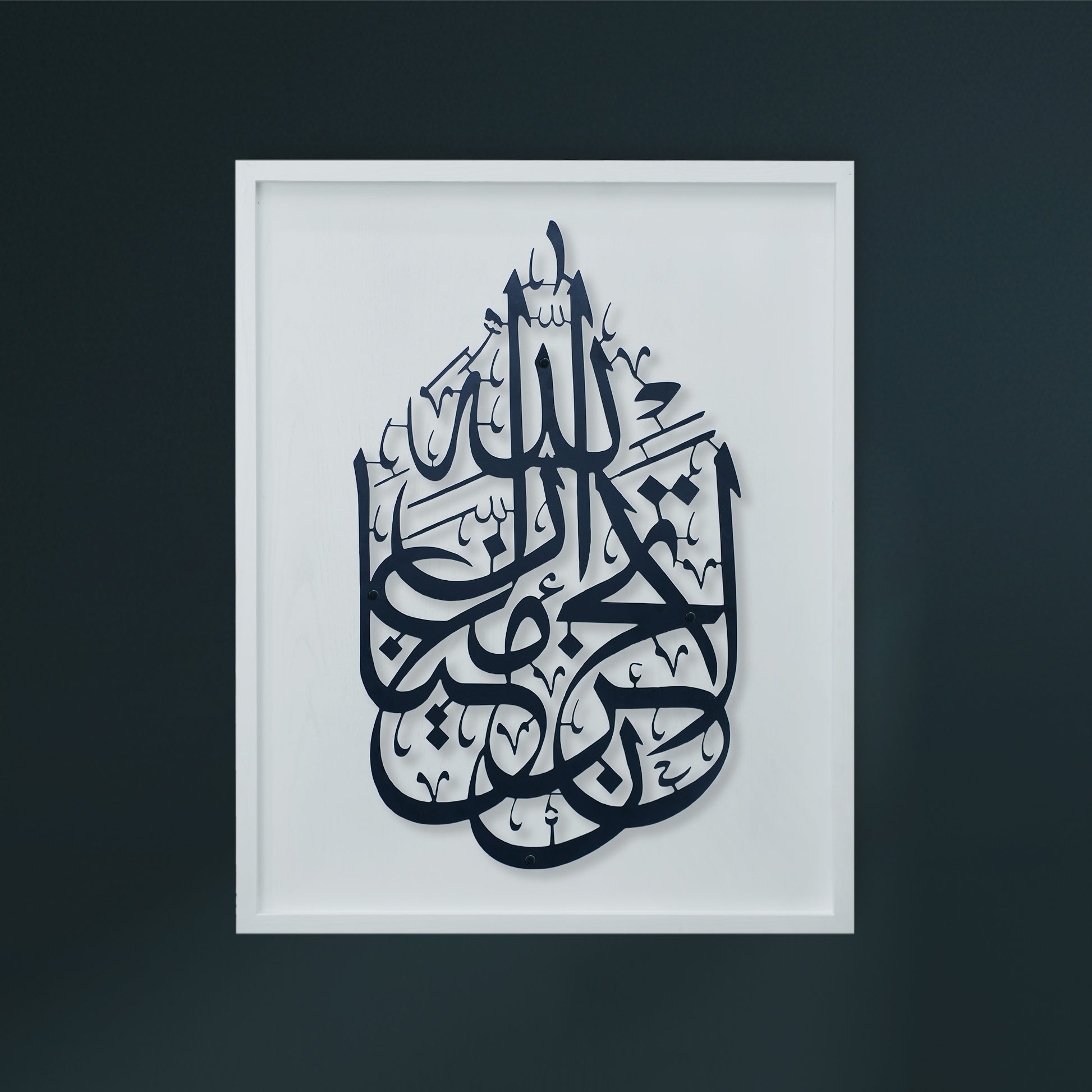 Framed Islamic calligraphy metal wall art featuring a black Arabic script design on a white wooden frame, enclosed in a simple white frame and displayed against a dark gray wall.