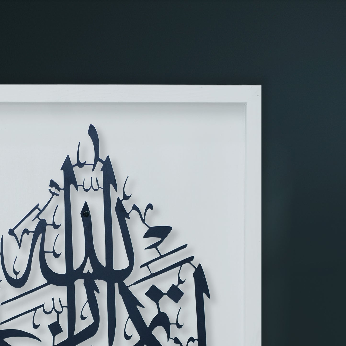 A close-up of framed metal wall art featuring Black Arabic calligraphy on a white Wooden frame, set against a dark charcoal wall.