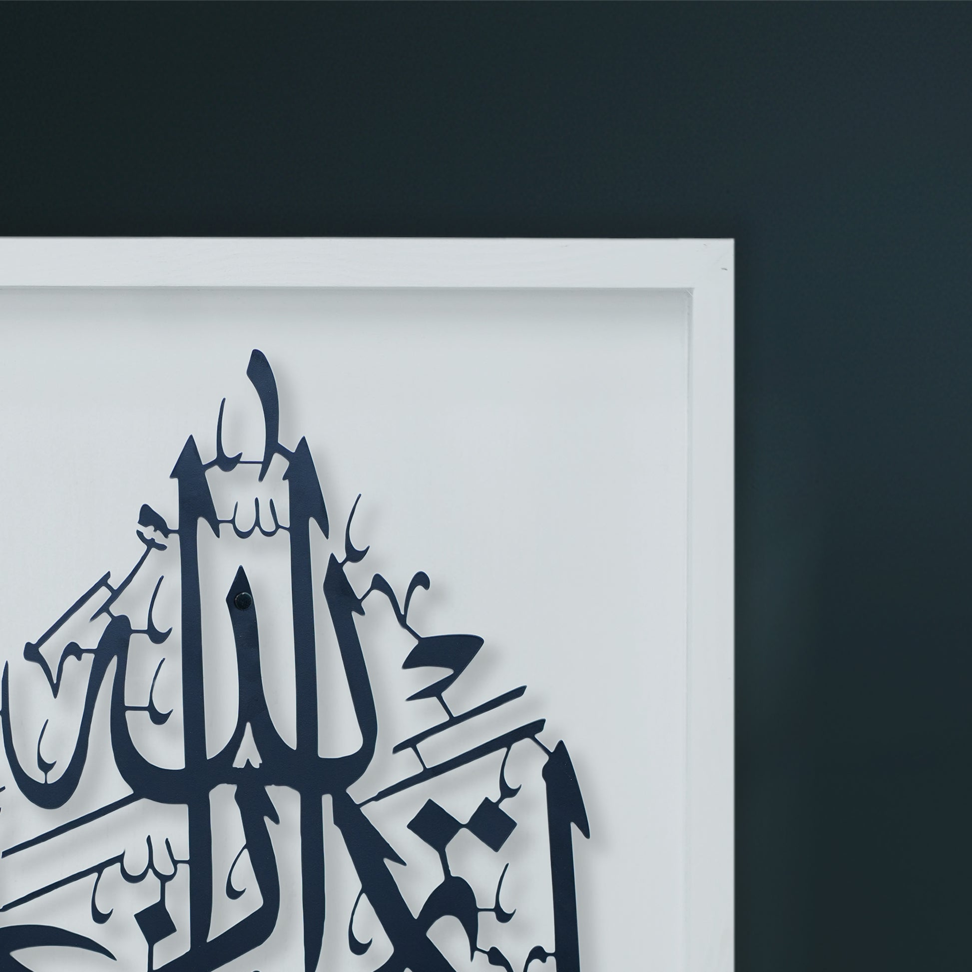 A close-up of framed metal wall art featuring Black Arabic calligraphy on a white Wooden frame, set against a dark charcoal wall.