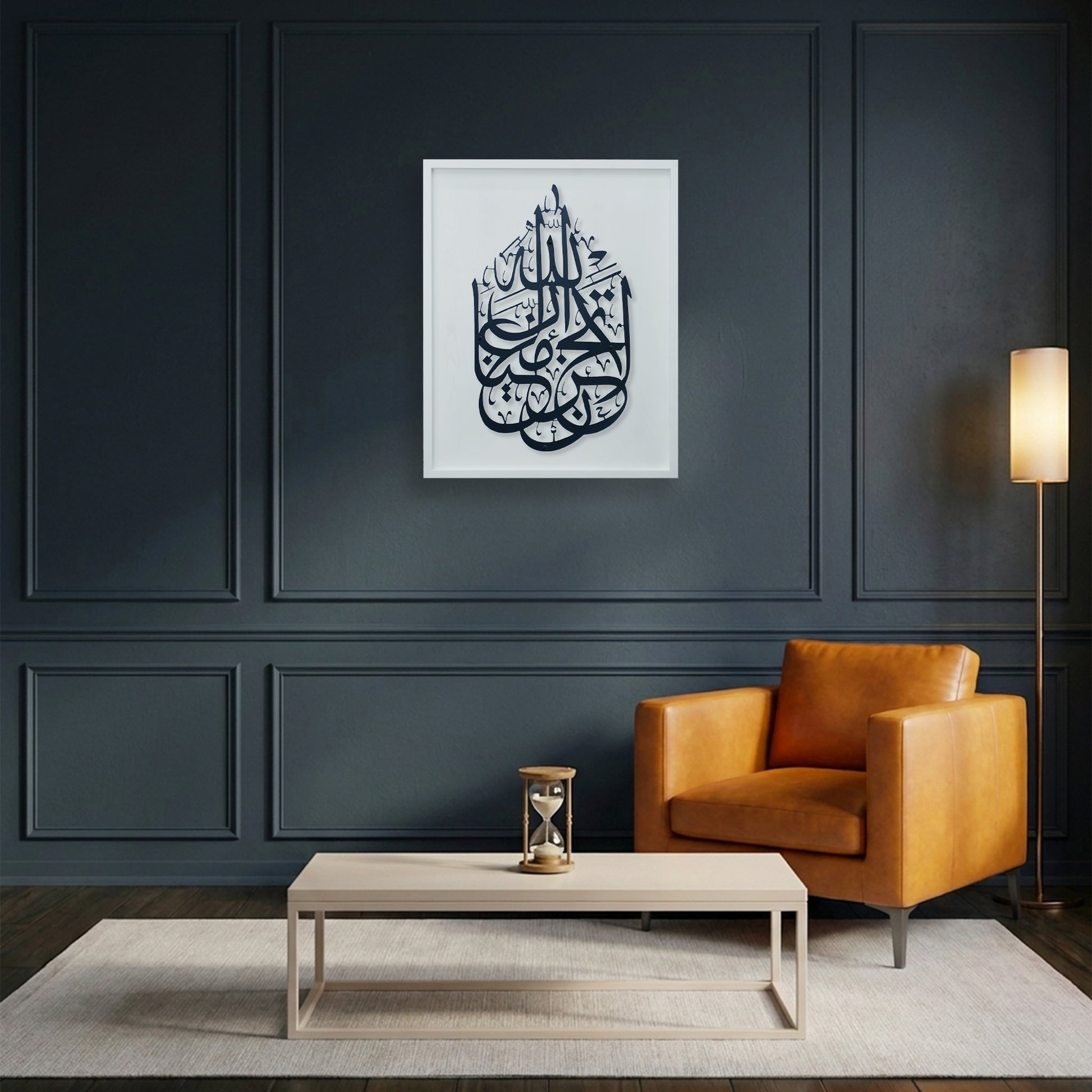 A framed black Arabic calligraphy metal wall art hangs on a dark navy paneled wall above a light wood coffee table, next to an orange leather armchair and a floor lamp.