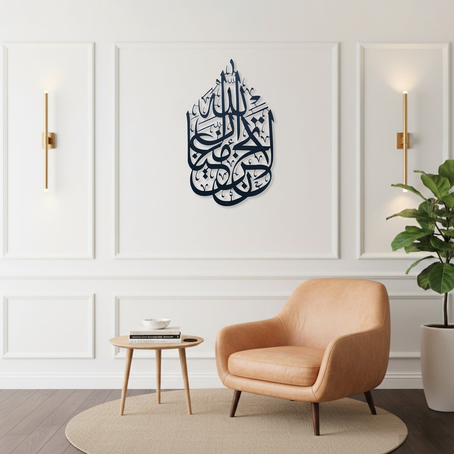 A styled interior room with a peach armchair, small round side table, potted plant, and wall sconces. The focal product appears to be the wall art: a dark black Arabic calligraphy La Tahzan İnnallahe Maana, Metal Wall Art piece mounted in the center of a white paneled wall.