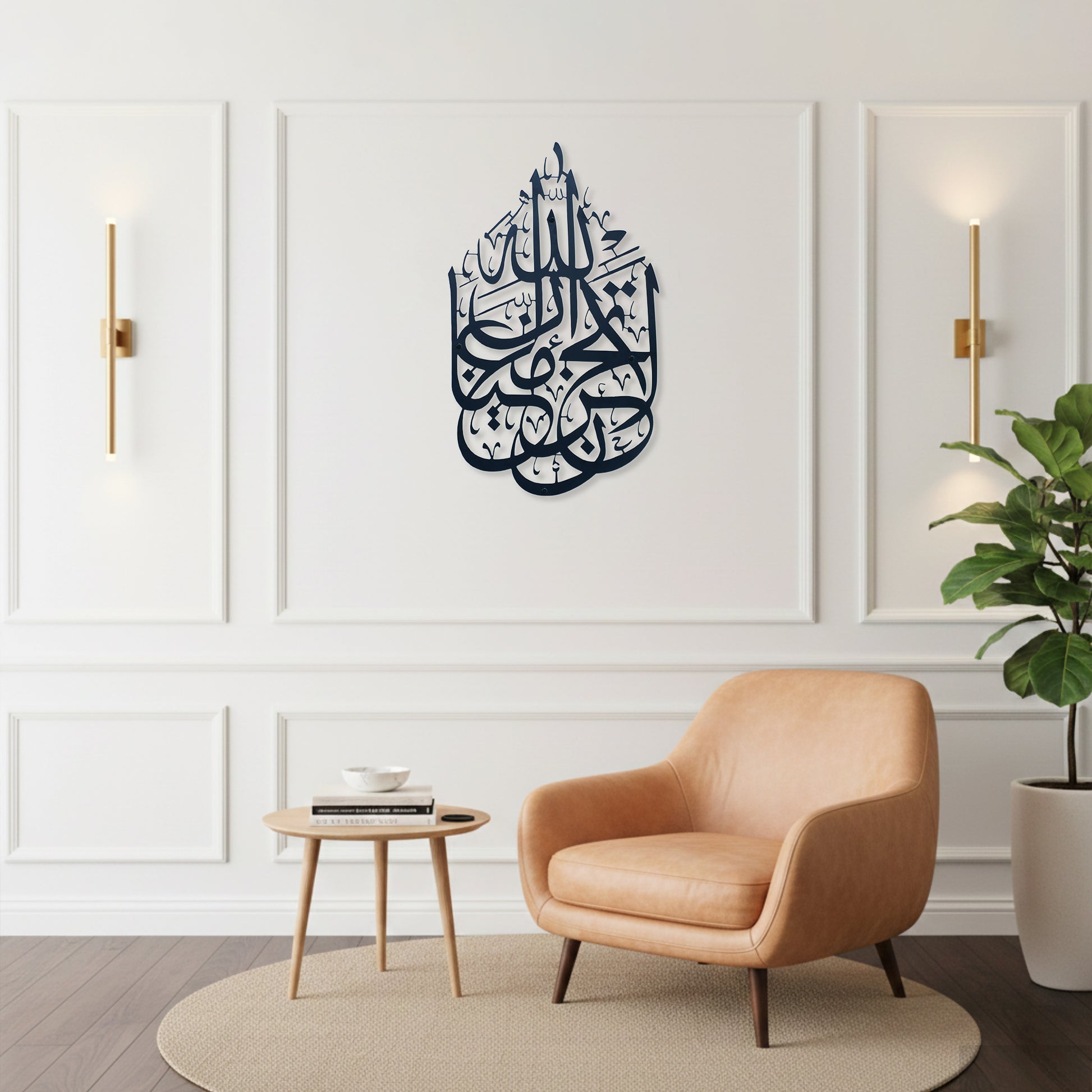 A styled interior room with a peach armchair, small round side table, potted plant, and wall sconces. The focal product appears to be the wall art: a dark black Arabic calligraphy La Tahzan İnnallahe Maana, Metal Wall Art piece mounted in the center of a white paneled wall.