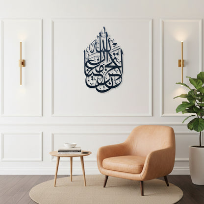 A styled interior room with a peach armchair, small round side table, potted plant, and wall sconces. The focal product appears to be the wall art: a dark black Arabic calligraphy La Tahzan İnnallahe Maana, Metal Wall Art piece mounted in the center of a white paneled wall.
