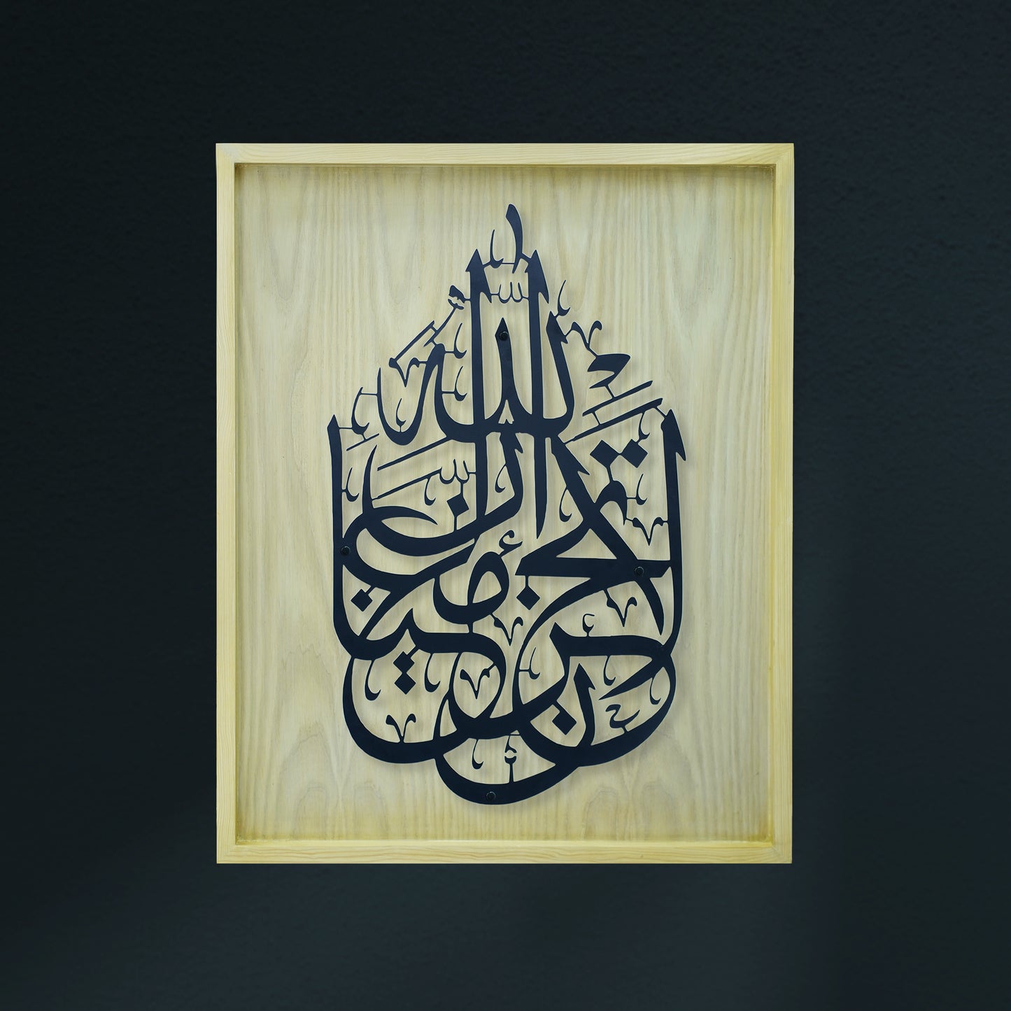 Black Arabic calligraphy art mounted on a light natural wood frame, set against a dark charcoal wall.