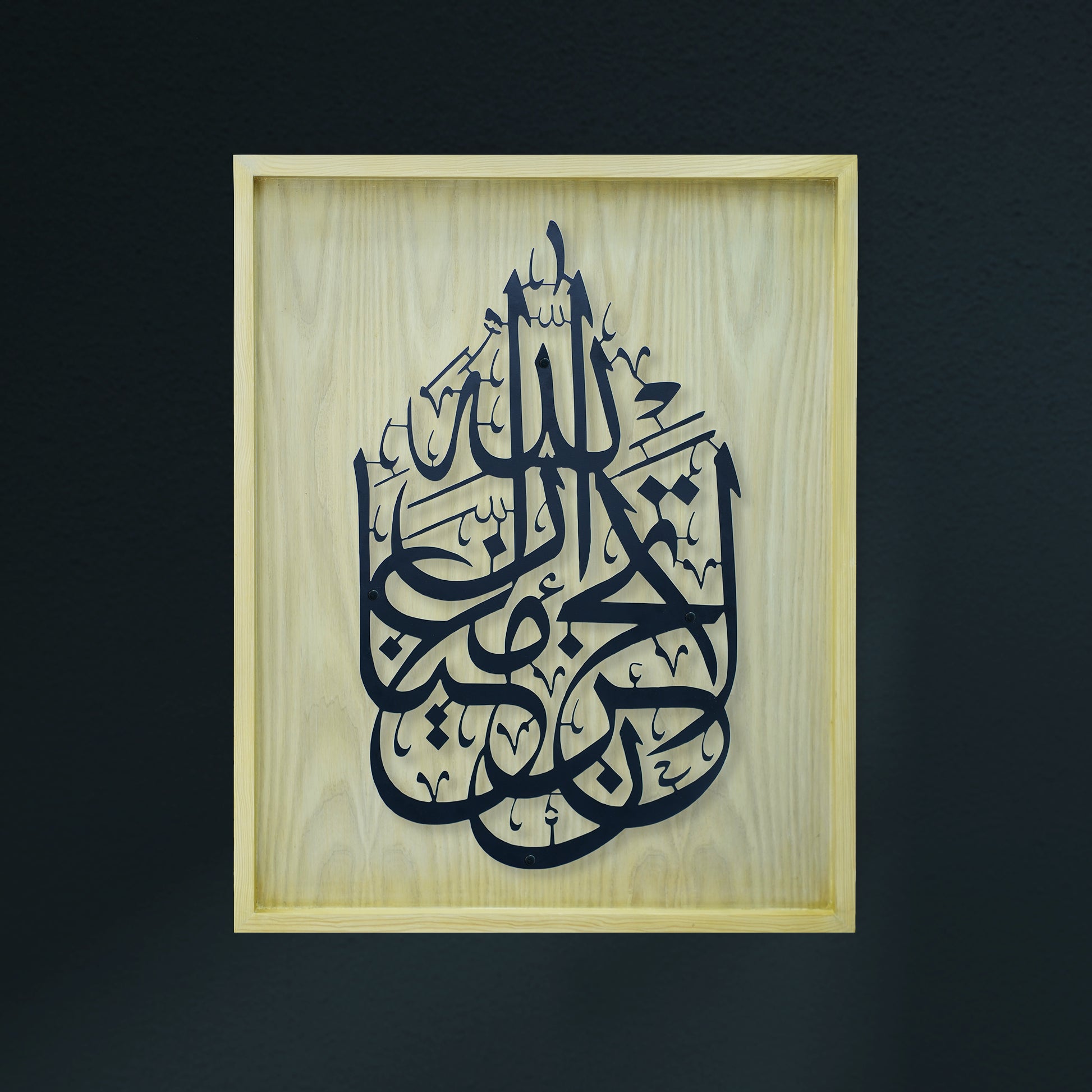Black Arabic calligraphy art mounted on a light natural wood frame, set against a dark charcoal wall.