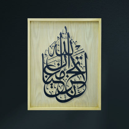 Black Arabic calligraphy art mounted on a light natural wood frame, set against a dark charcoal wall.