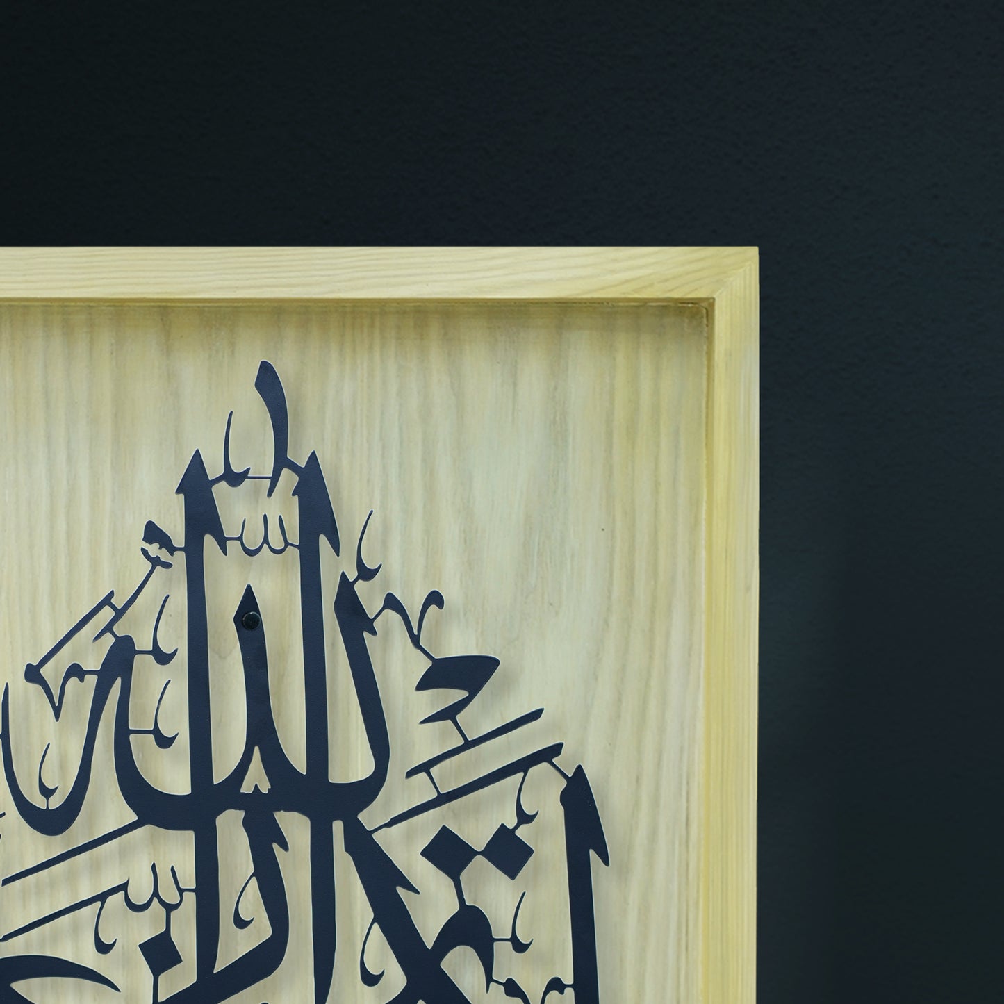 A close-up of framed wall art featuring black Arabic calligraphy on a light natural wood panel, set against a dark charcoal wall.