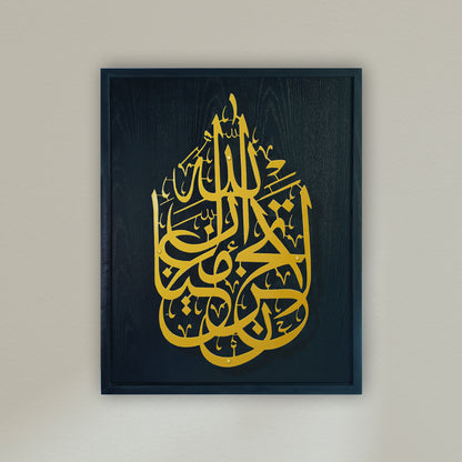 Framed Islamic wall art featuring intricate gold Arabic calligraphy on a blue wooden frame, mounted on a light beige wall.