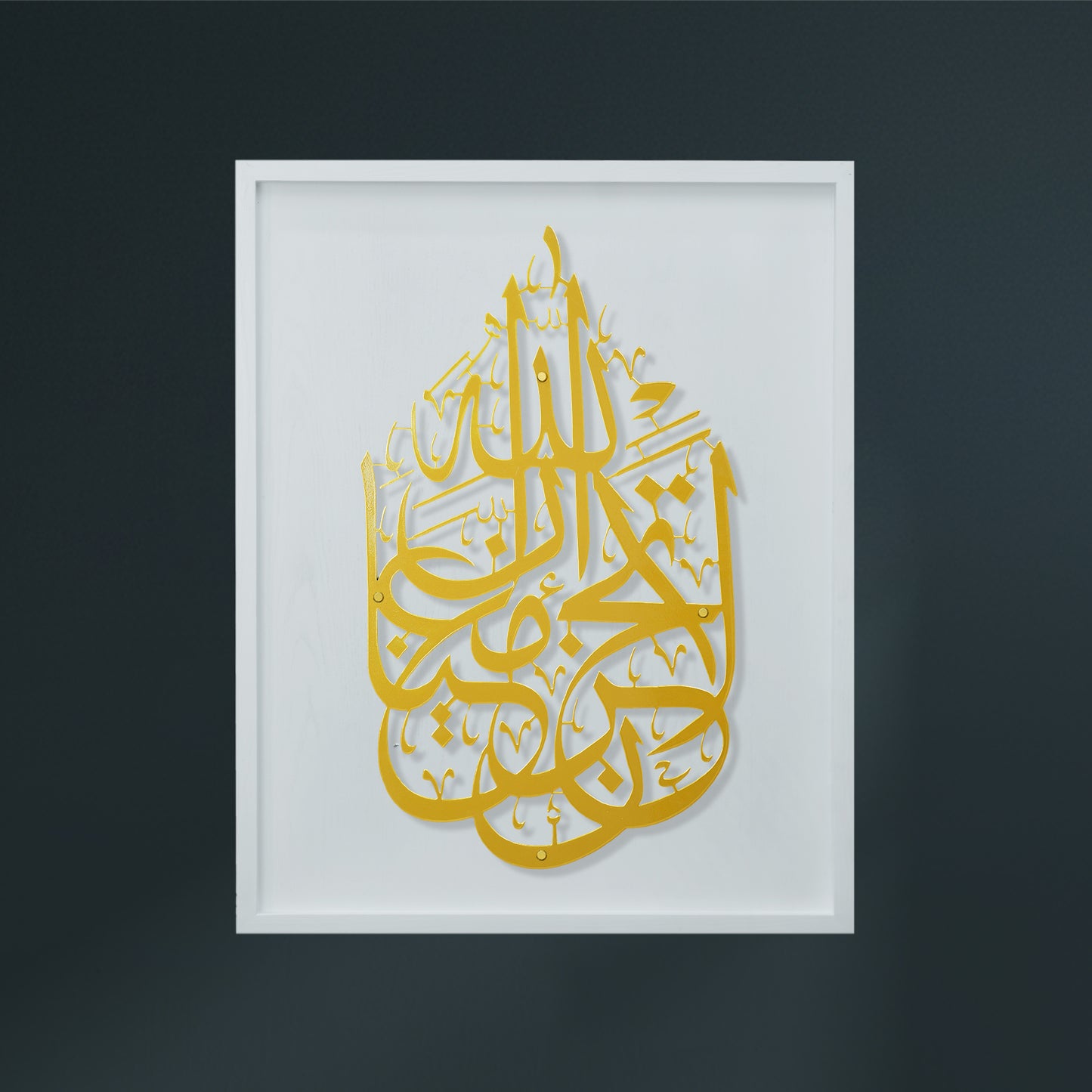 Gold Arabic calligraphy wall art mounted on a white square panel, displayed in a white frame against a dark gray wall.