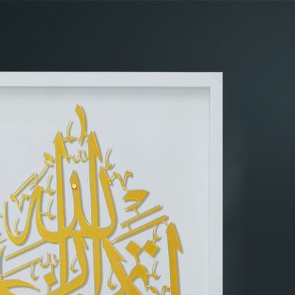 A close-up of a framed wall art piece featuring gold Arabic calligraphy on a white background, set against a dark charcoal wall.