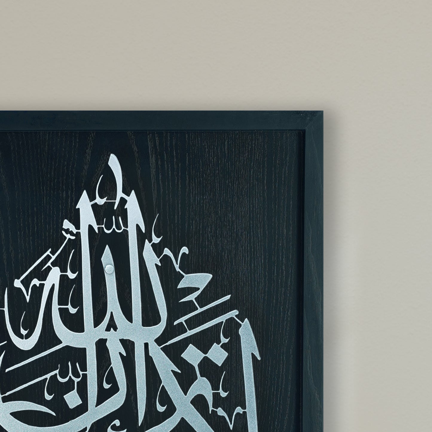 Black and Silver Framed Islamic calligraphy wall art featuring silver Arabic script on a Black wood-textured frame, shown hanging on a light beige wall.