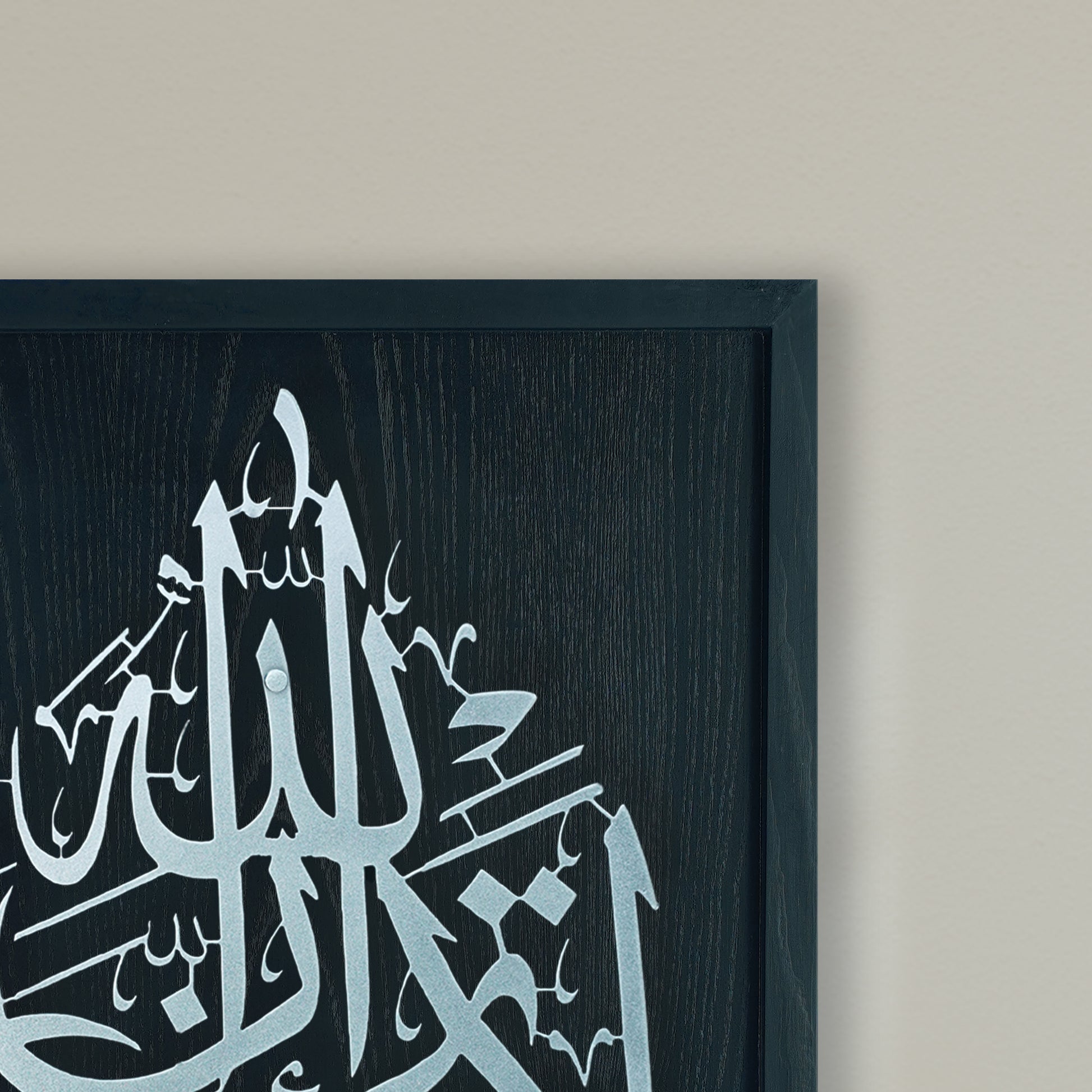 Black and Silver Framed Islamic calligraphy wall art featuring silver Arabic script on a Black wood-textured frame, shown hanging on a light beige wall.